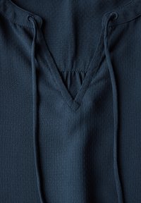Navyblauwe, textuurstof met een V-halslijn en trekkoordjes. Bevat kleine perforaties, die detail en ademend vermogen toevoegen.