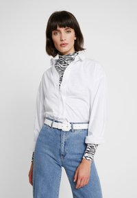 Hvid, langærmet skjorte over en sort-hvid zebra print turtleneck, parret med blå højtaljede jeans og et hvidt bælte.