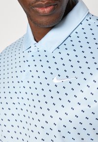 Ljusblå polo skjorta med en texturerad mönster av små marinblå rutor. Innehåller en kontrasterande vit Nike-logotyp på bröstet.