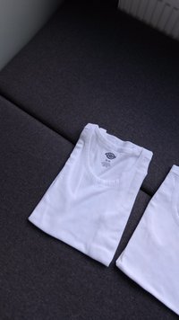 Duas t-shirts brancas dobradas sobre uma superfície cinza escura. As camisetas apresentam decotes redondos e uma textura de algodão suave.