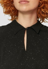 Schwarze Bluse mit einem subtilen Glitzer-Muster, ausgestattet mit einem kragenförmigen Ausschnitt und einem Schlüsselaugenverschluss mit Knopf. Weicher, strukturierter Stoff.