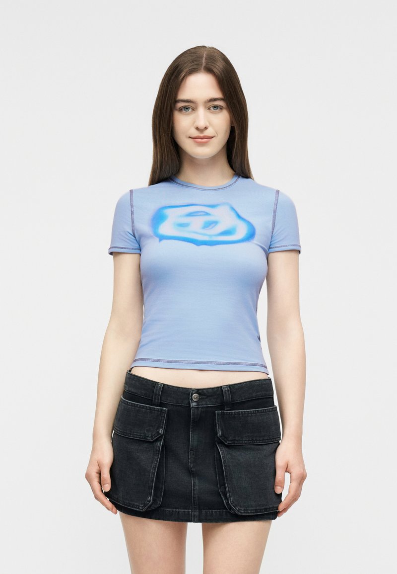 Νεαρή γυναίκα με μακριά καστανά μαλλιά που φοράει ανοιχτό μπλε εφαρμοστό τοπ με κοντά μανίκια και αφηρημένο σχέδιο, καθώς και μαύρη μίνι φούστα με μεγάλες μπροστινές τσέπες.