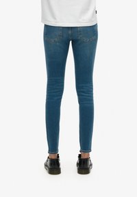 Jean slim bleu avec une texture lisse, présentant une taille mi-haute, des poches arrière et une coupe au niveau des chevilles, associée à des chaussures noires.