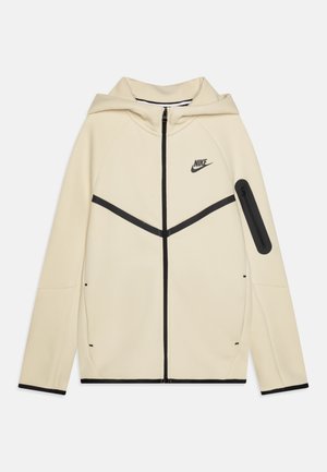 Nike mikina s kapucňou, krémová tkanina s čiernymi akcentmi, predný zips, čierny horizontálny pruhový dizajn, zipsový vrecko na rukáve, hladká textura.