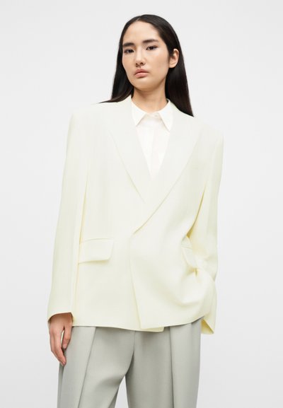 Δίδυμο σακάκι υπερμεγέθες σε απαλό κίτρινο ύφασμα, με αιχμηλές πέτες, δύο μπροστινές τσέπες και άνετη εφαρμογή. Φορεμένο πάνω από ένα ελαφρύ πουκάμισο.