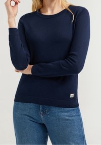 Kvinna i en enfärgad marinblå långärmad tröja och blå jeans, stående med armarna i kors, etikett som säger "Polo Club."