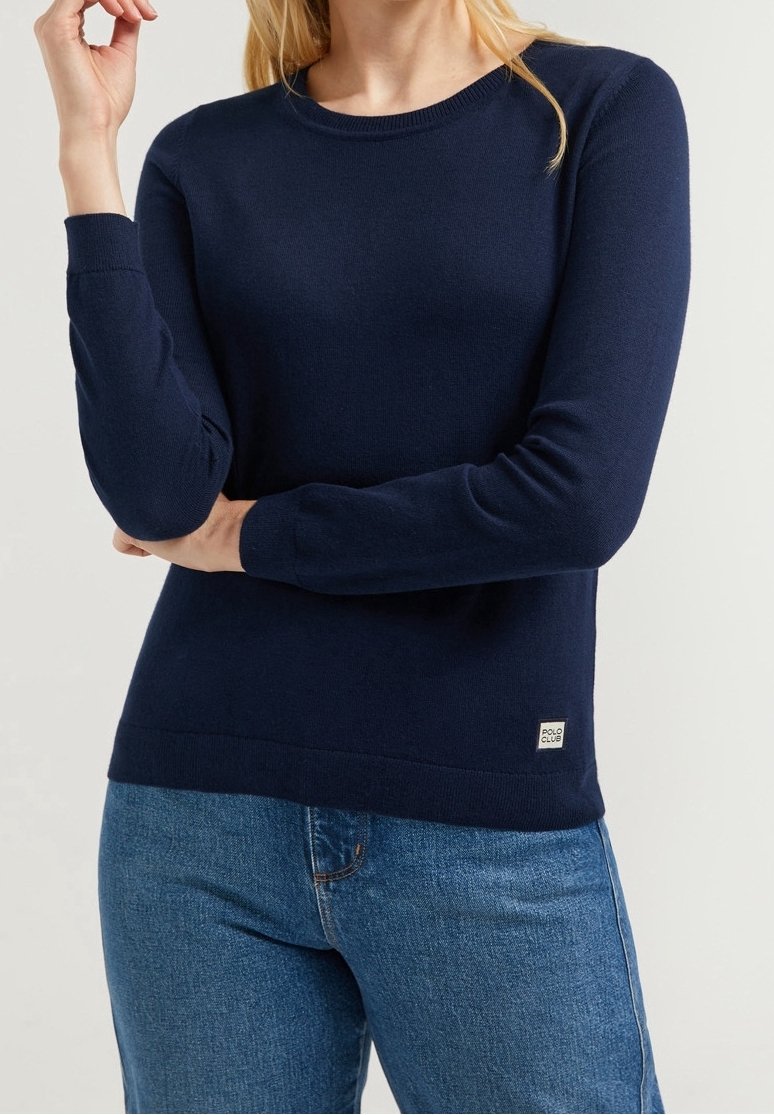 Kvinna i en enfärgad marinblå långärmad tröja och blå jeans, stående med armarna i kors, etikett som säger "Polo Club."