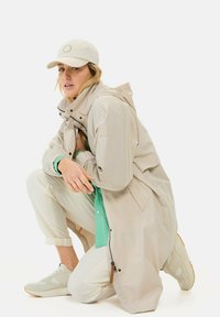 Hellbeige Regenjacke mit Kapuze, ausgestattet mit einem Reißverschluss und Fronttaschen. Trägt eine türkisfarbene Bluse und hellfarbige Hosen mit hellen Schuhen.