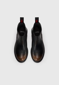 Une paire de bottines Chelsea en cuir noir avec des bouts marron, des semelles noires et des languettes rouges étiquetées "DAMPER" orientées vers le haut sur fond blanc.
