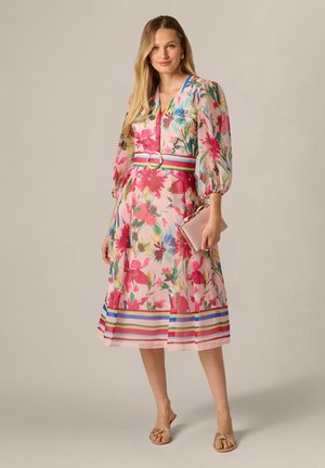 Kobieta w kwiecistej sukience midi z bufiastymi rękawami, w paski w talii, trzymająca różową kopertówkę, stojąca na jasnobeżowym tle.