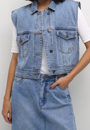 Person trägt ärmellose hellblaue Jeansjacke über weißem kurzärmligem Hemd und passend hohem Jeansrock, Hand in der Tasche.