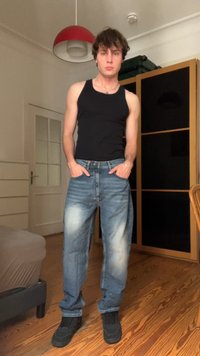 Camisola preta, jeans azul desbotado com um ajuste descontraído e ténis pretos. O ambiente apresenta um piso de madeira e uma luz pendente vermelha.
