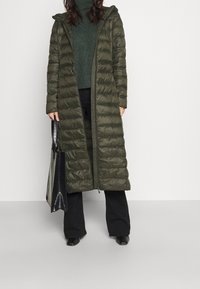 Długi, oliwkowozielony puchowy płaszcz z kapturem, pikowany, z pełnym zamkiem, w połączeniu z ciemnozielonym swetrem i czarnymi spodniami.
