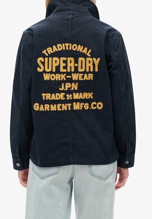 Personne portant une veste bleu marine foncé avec un texte jaune brodé "Traditional Super-Dry Work-Wear J.P.N Trade Mark Garment Mfg.Co" dans le dos.