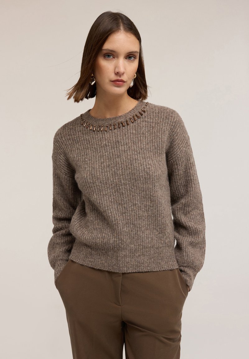 Maglione marrone a coste con scollo rotondo e decorazioni in perline attorno al colletto. Il modello indossa pantaloni marroni a sigaretta. Tessuto morbido.