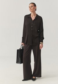 Sort langærmet skjorte med knapper og vide bukser, parret med sorte flade sko. Outfittet har en glat tekstur og en afslappet pasform.
