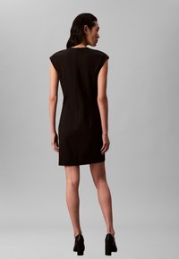 Sort ermeløs kjole med strukturerte skuldre, knelengde snitt, glidelåsdetalj bak, supplert med buede sømmer og tettsittende design.