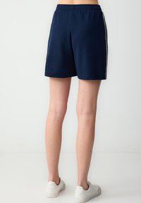 Shorts sportivi blu navy in tessuto testurizzato, con vita elastica e strisce bianche sui lati. Modello che indossa scarpe da ginnastica bianche.