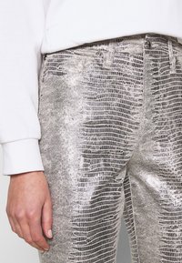 Silverfärgade byxor med ormskinnsmönster och en texturerad yta, med en figurnära design och synliga sömmar. Bärs tillsammans med en vit sweatshirt med uppvikta ärmar.