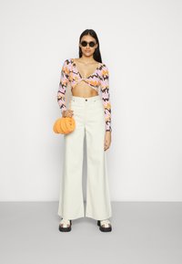 Top cropped de manga longa colorido com padrões de ondas em rosa, laranja e castanho; calças largas creme; bolsa redonda de palha laranja; sandálias de plataforma pretas.