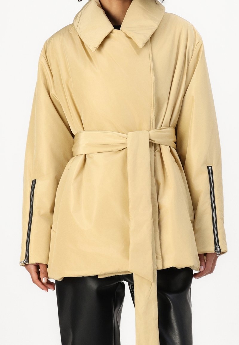 Doudoune beige ceinturée avec col oversized et détails de fermeture éclair noirs sur les manches, portée avec un pantalon en cuir noir.
