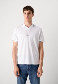 Biele polo tričko s textúrovaným dizajnom, s čiernym golierom a pruhovanými akcentmi. Logo zobrazené na hrudi. Spojené s modrými džínami.