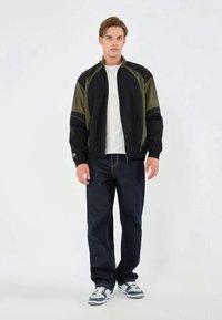 Joven de pie con chaqueta negra y oliva, camiseta blanca, jeans oscuros y zapatillas blancas, contra un fondo claro y sencillo.