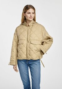 Beżowa pikowana kurtka z kapturem, wyposażona w dwie przednie kieszenie, zapięcie na zatrzask oraz regulowane sznurki na dole. Idealna na codzienne noszenie.