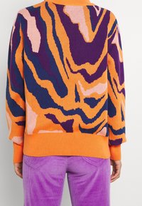 Suéter de punto en naranja, púrpura y rosa con un patrón de olas; cuenta con puños y dobladillo acanalados. Combinado con pantalones de pana morados.