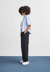 Chemise à manches courtes bleu clair, pantalon noir taille haute avec poches, baskets blanches et lunettes de soleil foncées. Tissu lisse et coupe décontractée.