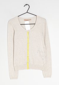 Ljusbeige cardigan med rund hals, gul knappslå, ribbstickade muddar och fåll samt dekorativa silverdetaljer på ena sidan.