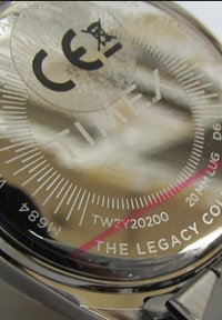 Sølvfarvet metalur med en skinnende overflade. Viser "TIME.X" og "THE LEGACY COL." samt CE-mærkning, model og lugmål.