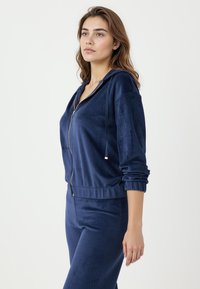 Marineblauwe zip-up hoodie van zachte, fluweelachtige stof met een ontspannen pasvorm, zakken aan de voorkant en een verstelbare trekkoord bij de capuchon.