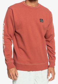 Crewneck sweatshirt i bränd orange, tillverkad av mjuk bomull. Har en liten logotyp-patch och vit branding längs vänster ärm.
