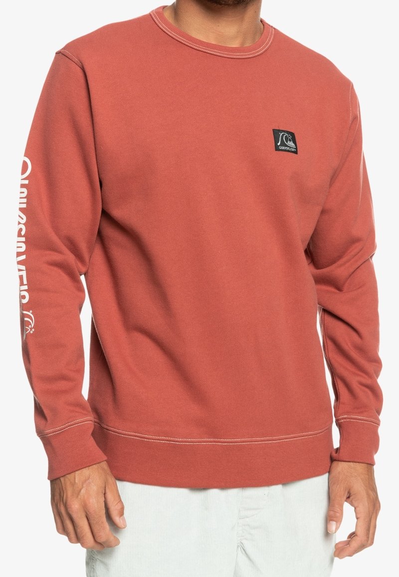 Crewneck sweatshirt i bränd orange, tillverkad av mjuk bomull. Har en liten logotyp-patch och vit branding längs vänster ärm.