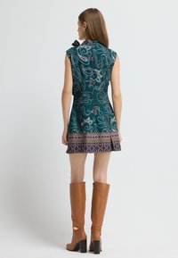 Kurzärmliges türkisfarbenes Kleid mit wirbelnden Mustern, einem schwingenden Rock und einem dekorativen Band. Kombiniert mit hohen braunen Lederstiefeln.