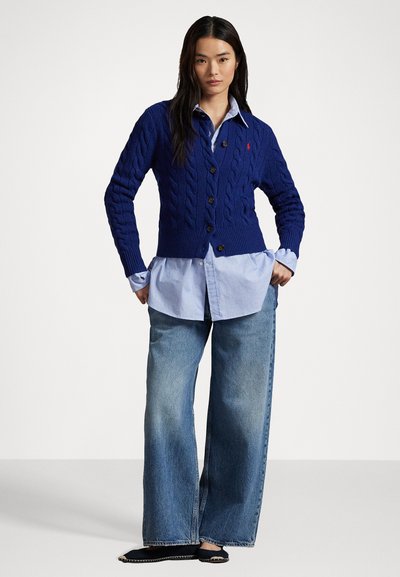 Marineblå kabelstrikket cardigan med V-hals, brune knapper og ribbestrikket kant, lagt over en lyseblå skjorte med knapper og vide jeans.