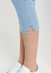 Jeans capri en denim bleu clair avec un ourlet cropped présentant une petite fente, associés à des baskets blanches. Détails de couture le long des bords.