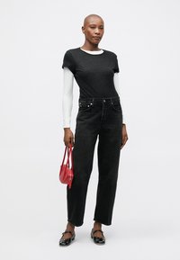 Cilvēks ar īsiem matiem, ģērbts slāņos melnos un baltos garajās piedurknēs, melnās džinsās, melnās plakanās kurpēs, tur nelielu sarkanu rokassomu, stāv uz baltā fona.