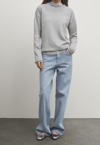 Sweat-shirt gris à col montant, coupe ample et poignets côtelés. Associé à un jean large bleu clair et des chaussures argentées à enfiler.