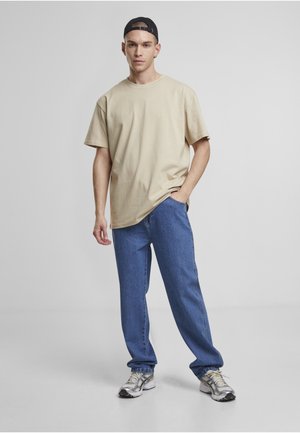 Jeune homme debout portant un t-shirt beige, un jean bleu, des baskets grises et une casquette de baseball noire à l’envers sur un fond gris uni.