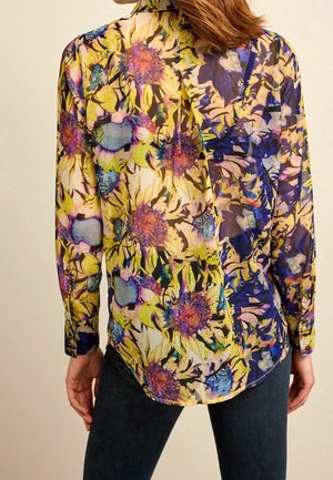 Donna che indossa una camicetta trasparente a maniche lunghe con una vivace stampa floreale nei toni del giallo, viola e blu, abbinata a jeans scuri.