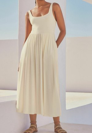 Vrouw die een mouwloze crèmekleurige midi-jurk draagt met een nauwsluitend lijfje en een geplooide rok, staand tegen een lichtblauwe en witte architecturale achtergrond.