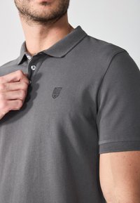Siva polo majica od teksturirane tkanine, s trostrukim zakopčavanjem i izvezenim logotipom na lijevoj strani prsiju.