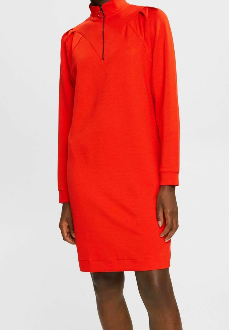 Czerwona sukienka w stylu shift z długimi rękawami, wysokim kołnierzem i zapięciem na zamek. Posiada marszczone detale przy ramionach oraz gładki, elastyczny materiał.