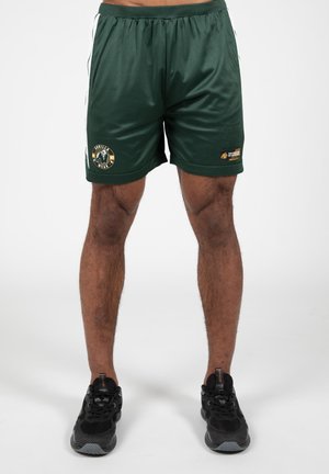 Groene sportieve shorts van gladde stof, met een logo-opdruk en witte zijstrepen. Gedragen met zwarte sneakers.