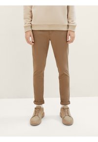 Dolní část těla osoby v béžových nohavicích s ohrnutými nohavicemi, bílých ponožkách, béžových teniskách a béžovém mikině na jednobarevném pozadí.