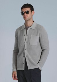 Gray ribbed cardigan with a collared neckline, button-down front, and a chest pocket. Worn over a light gray shirt. Textured fabric.