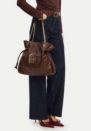 Mujer con vaqueros oscuros y camiseta marrón, sosteniendo un gran bolso de cuero marrón con tirantes de cadena dorada y detalle del logo.