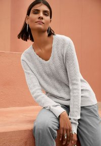 Frau mit dunklen Haaren, die einen gemusterten weißen Pullover, graue Hosen und hängende Ohrringe trägt, sitzt an einer pfirsichfarbenen Wand.